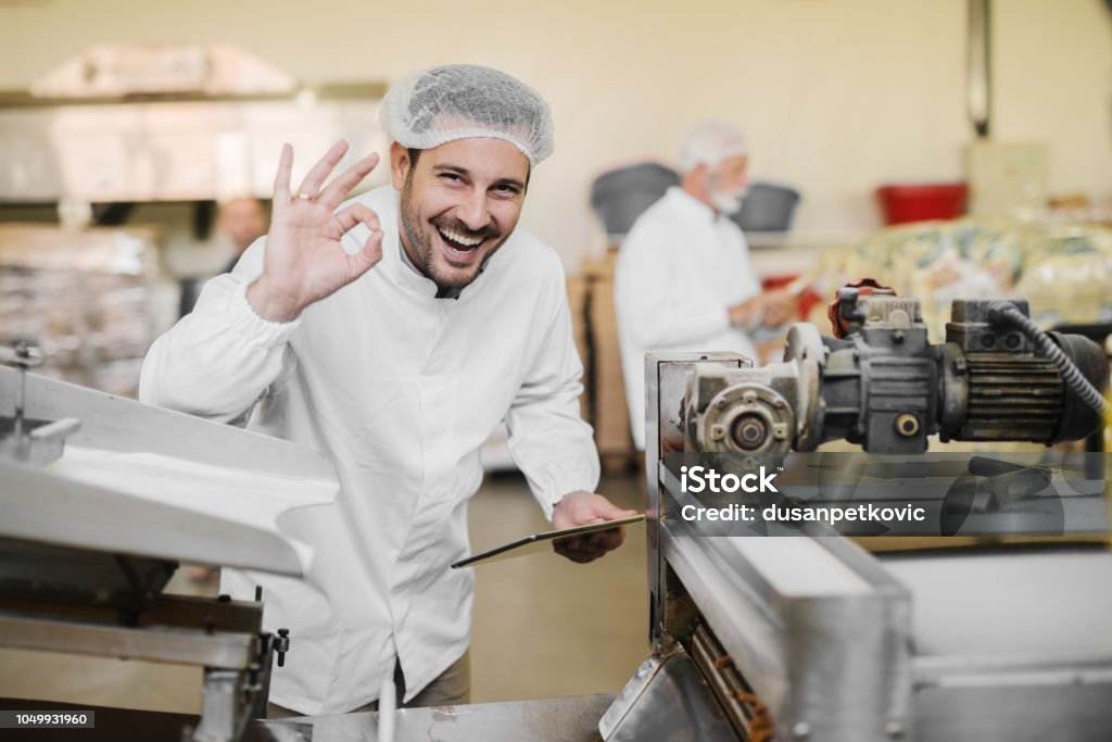 良い品質。食品工場で無菌服陽気な笑みを浮かべて若い男の写真。1 つの手と他のことを身振りで示す製品の品質でタブレットを保持することをお勧めします。 - 食べ物のロイヤリティフリーストックフォト