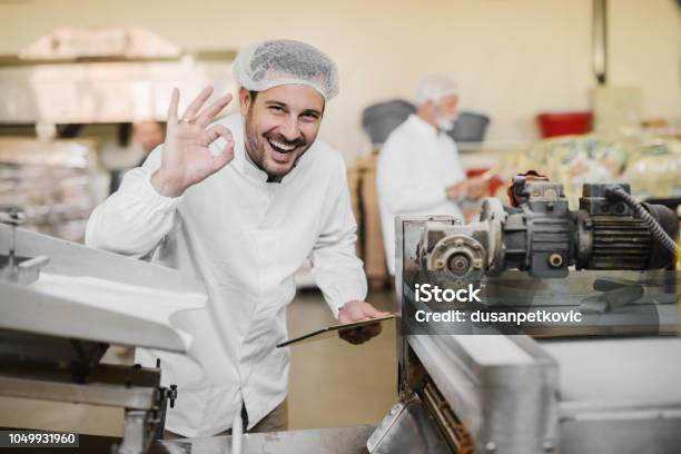 Good Quality Picture Of Cheerful Smiling Young Man In Sterile Clothes In Food Factory Holding Tablet In One Hand And With Other Gesturing That The Quality Of Product Is Good Stock Photo - Download Image Now