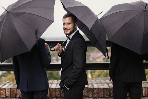 Stylish groom and groomsmen standing under black umbrella and posing. Confident man in suit holding umbrella in rainy outdoors. Rich Businessman. Finance wealth and business