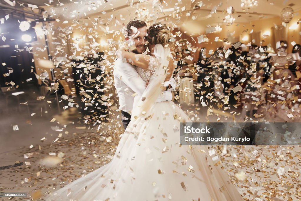 Gorgeous bride and stylish groom dancing under golden confetti at wedding reception. Happy wedding couple performing first dance in restaurant. Romantic moments Wedding Stock Photo