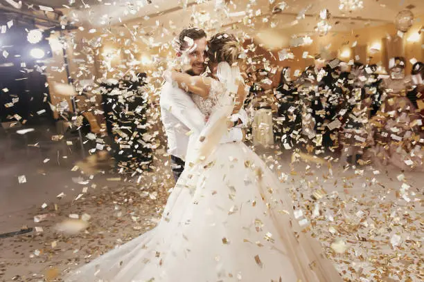 Photo of Gorgeous bride and stylish groom dancing under golden confetti at wedding reception. Happy wedding couple performing first dance in restaurant. Romantic moments