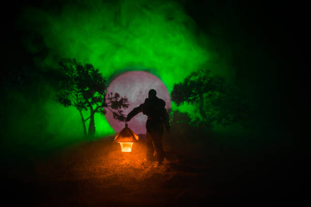 istock Man in raincoat coming from dark forest with glowing lantern in his hand concept. Silhouette of a horror killer with lamp 1050002906