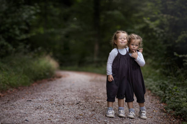 Two little identical twin girls in dark overalls standing on the forest road hugging and grimacing.