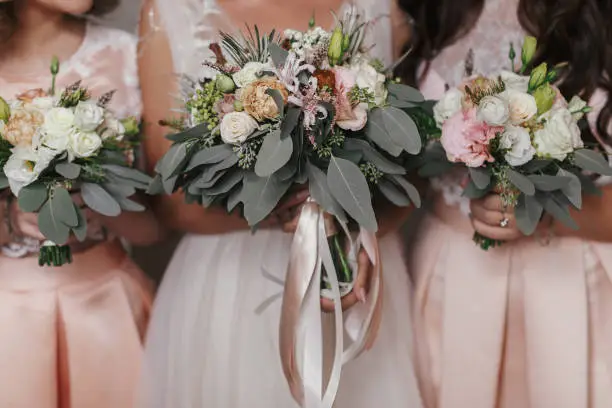 Bridesmaids and bride holding modern wedding bouquets of pink roses and green eucalyptus with pink ribbons. Stylish Contemporary bouquets on soft fabric. Wedding arrangements