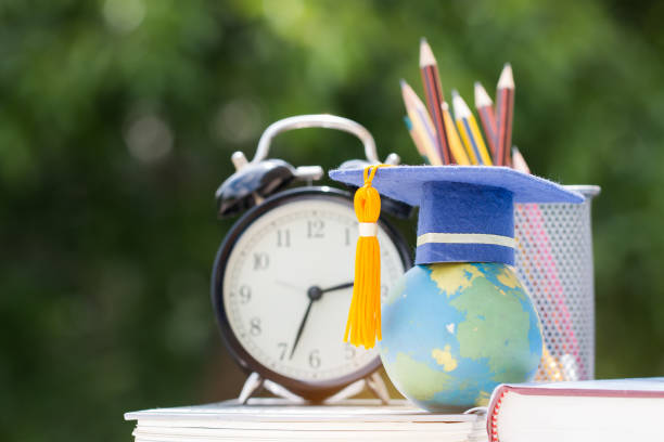 istock Graduate or Education knowledge study abroad concept : Graduation cap on textbook with pencil box and retro clock on desk, blur green light background. Back to School 1050111802