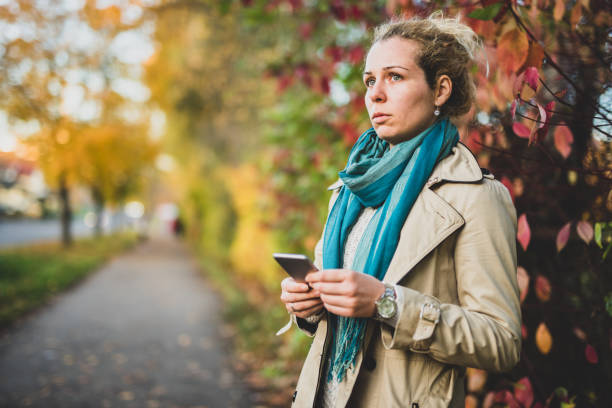 Beautiful woman holding smart phone and waiting for someone. Blondie female standing on street and looking in view. norway autumn oslo tree stock pictures, royalty-free photos & images