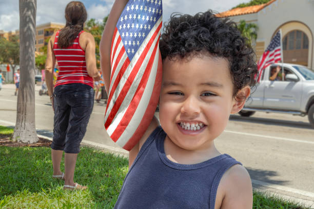 Young latina Mother with her happy boy at the parade. Fourth of July parade passes by as a Toddler boy holds the American flag up-looking at the camera with a-big smile. parade stock pictures, royalty-free photos & images