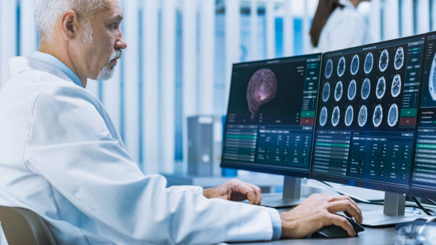 istock Experienced Senior Scientist Working with CT/ MRI Brain Scan Images on a Personal Computer in Laboratory. Neurologists / Neuroscientists in Research Center Work on Brain Tumor Cure. 1050311756
