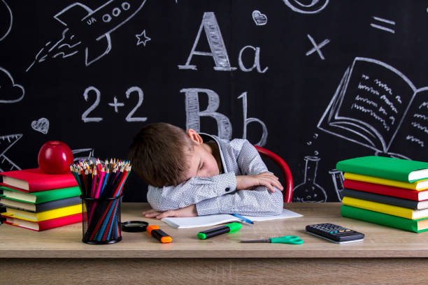 istock Schoolboy sleeping at the desk with a head leaned on the hands, surrounded with school supplies. Chalkboard as a background 1050312554