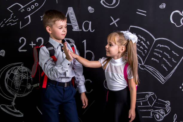 istock School mates standing before the chalkboard as a background with a backpack on their backs claping their hands looking to each other. Boy and girl 1050315668