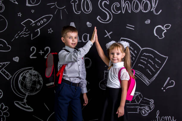 istock Boy and girl. School mates standing before the chalkboard as a background with a backpack on their backs claping their hands looking straight to the camera 1050315716