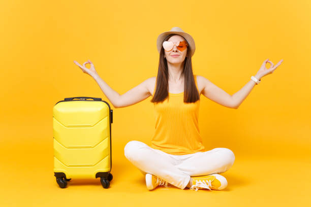 Traveler tourist woman in summer hat sit with suitcase crossed legs, meditate spread hands, isolated on yellow orange background. Passenger traveling abroad on weekends getaway. Air flight concept viajero turismo mujer sombrero de verano sentarse con maleta cruzó las piernas, meditar las manos propagación, aisladas sobre fondo amarillo naranja. pasajeros viajando en el extranjero en escapada de fin de semana. concepto de vuelo de aire.