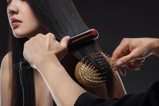 vista parcial del estilista del cabello alisar el cabello de la mujer con plancha de pelo y cepillo aislados en negro - straightener fotografías e imágenes de stock