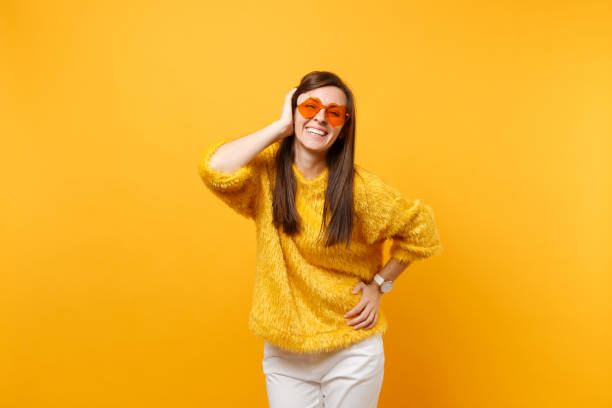 Smiling funny young woman in fur sweater, white pants and heart orange glasses keeping hand near ear isolated on bright yellow background. People sincere emotions, lifestyle concept. Advertising area sonriente mujer joven divertida en piel jersey, pantalón blanco y gafas de corazón naranja manteniendo la mano cerca de oído aislado sobre fondo amarillo brillante. emociones sinceras de personas, concepto de estilo de vida. Área de publicidad.