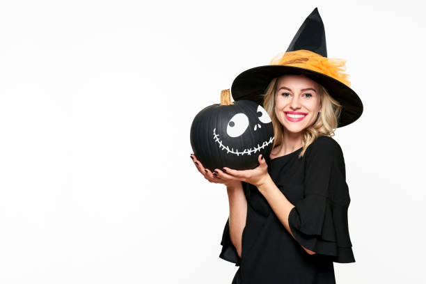 Gorgeous smiling Halloween Witch holding a Jack o Lantern. Beautiful young woman in witches hat and costume holding pumpkin over white background. stock photo
