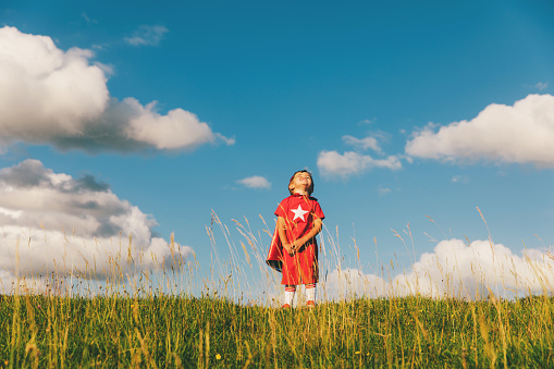 A young English boy dressed up in a red superhero costume with a white star stands in an English countryside grass field waiting to be super. The weather is pleasant with blue skies and green grass. The boy looking up with a confident smile and hope for being great. Image taken in Gloucestershire, United Kingdom.