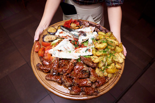 Cooked meat with vegetables on a large wooden board in the hands of the waiter.