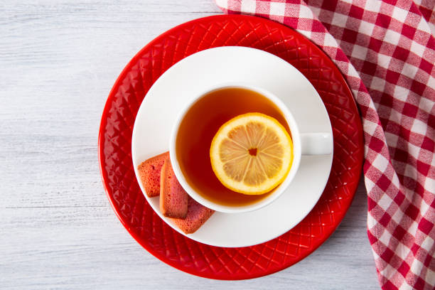 istock Tea in a white cup with lemon, three red cupcakes on a white plate. Napkin in a white and red cage. White wood background. Top view. Copy space. 1050445568