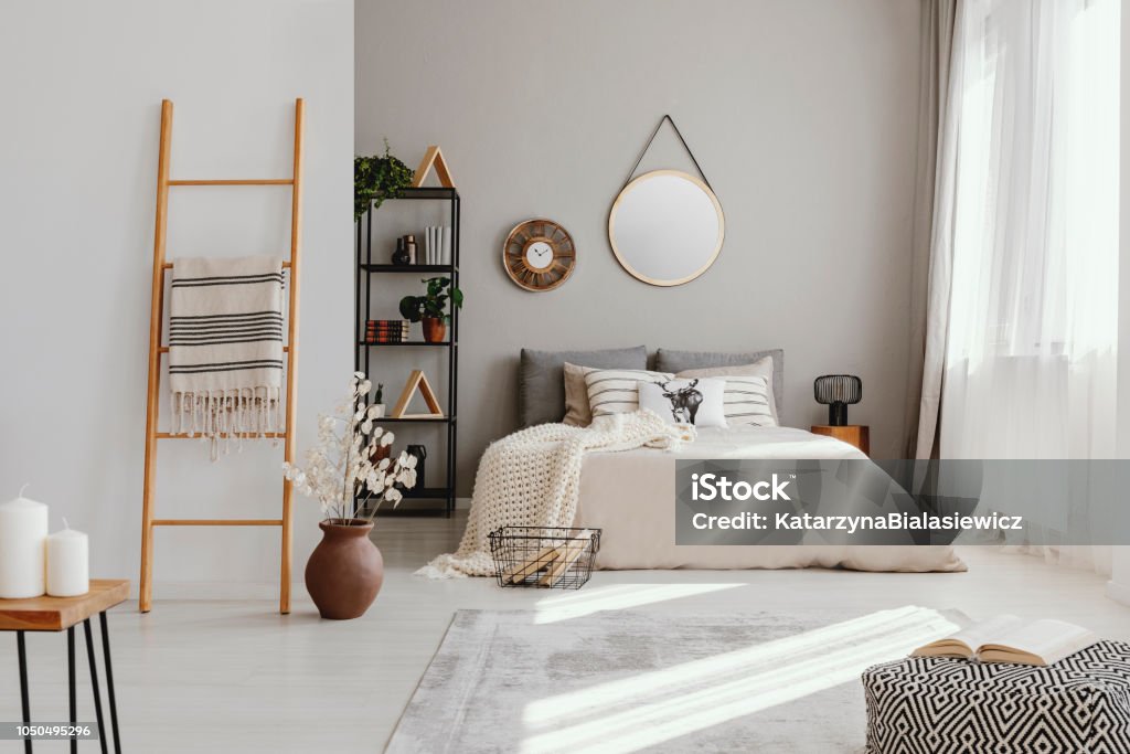 Espejo y reloj por encima de la cama en el interior de dormitorio brillante con PUF y flores junto a la escalera. Foto real - Foto de stock de Dormitorio - Habitación libre de derechos