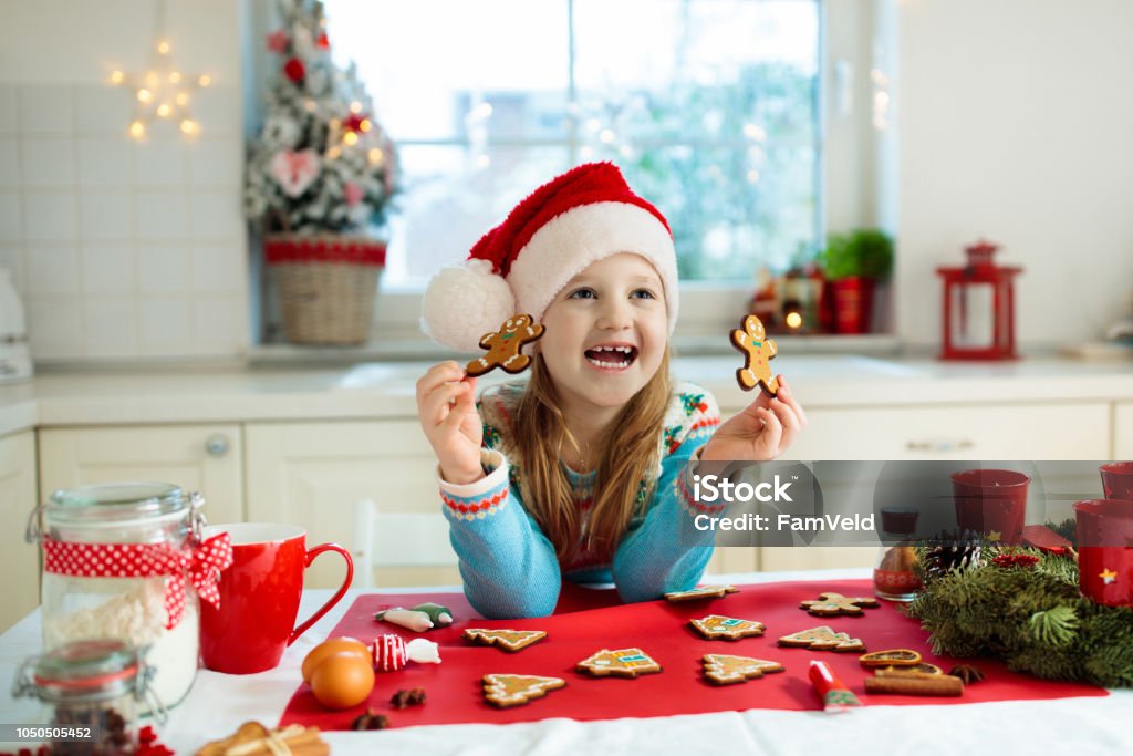 Kids bake Christmas cookies. Child cook for Xmas. Kids bake Christmas cookies. Child in Santa hat cooking, decorating gingerbread man for Xmas celebration. Family preparing sweets in white kitchen with Christmas tree on snowy winter day. Child Stock Photo