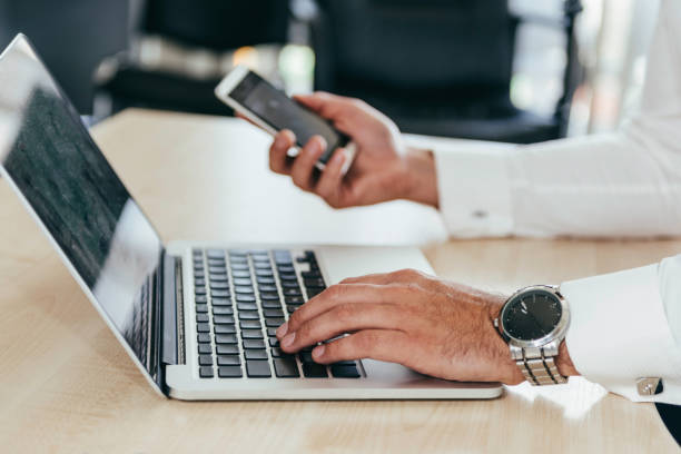 istock Midsection Of Businessman Using Laptop While Holding Mobile Phone On Table In Office 1050537256