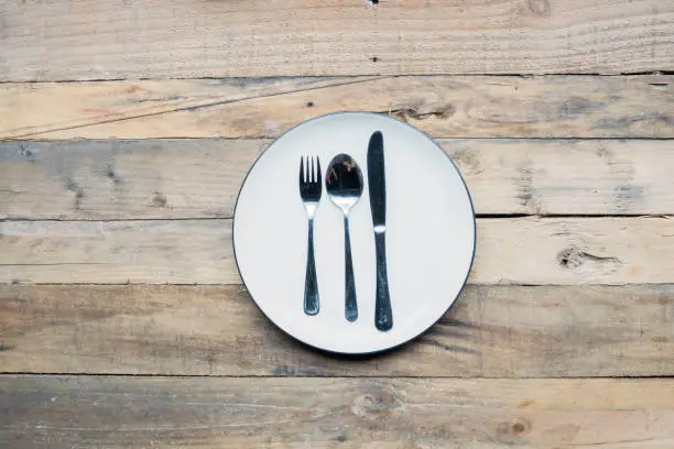 Closeup of a porcelain plate with steel spoon, fork and knife on the wooden table