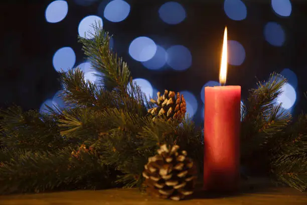 Image of a red Christmas candle with fir tree branches and pine cones on blurred sparkling light background