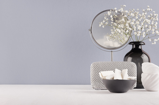 Perfect stylish decoration for home in grey colors - black glass vase with small fluffy flowers, mirror, female silver cosmetic bag, bowl sponges on soft ligth white wood table.