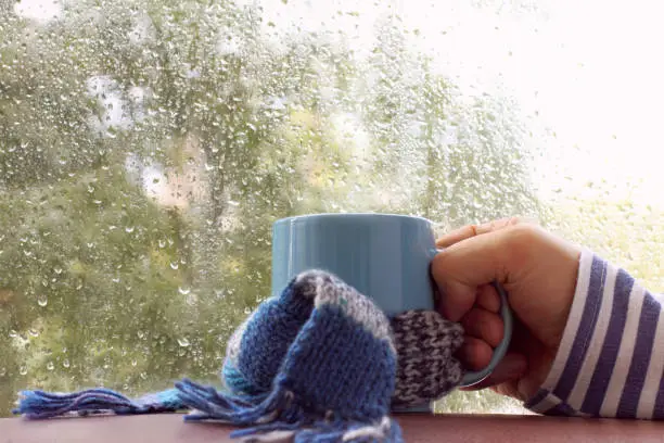 hand with cup on table, against the window with drops after rain