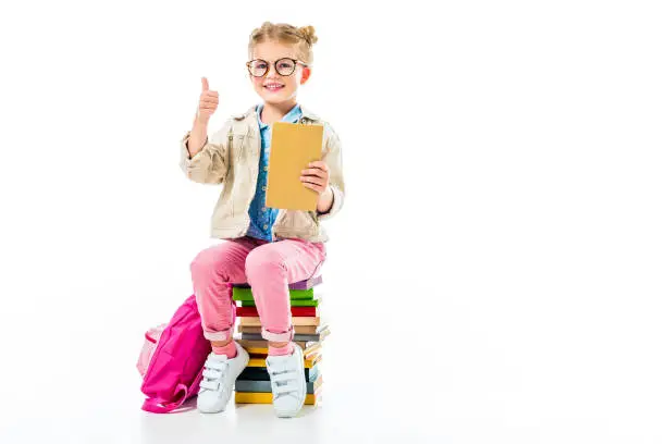 Photo of adorable smiling pupil showing thumb up while sitting on pile of books isolated on white, knowledge concept