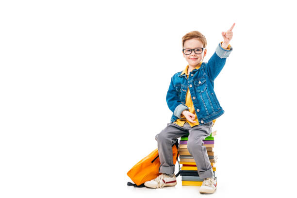 istock smiling schoolboy pointing up while sitting on pile of books with backpack, isolated on white 1050662730