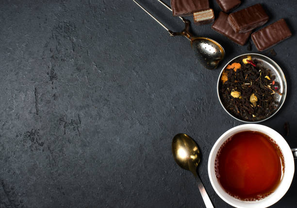Chocolates and black tea with herbs. Metal tea strainer, spoon. Dark background. Copy space. Flat lay