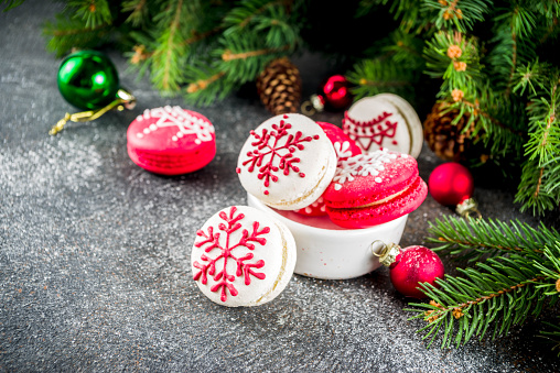 Creative idea for Christmas sweet treats, funny red and white macaron cookies decorated xmas ornament and snowflakes, dark rusty background with christmas tree branches and decorations, copy space