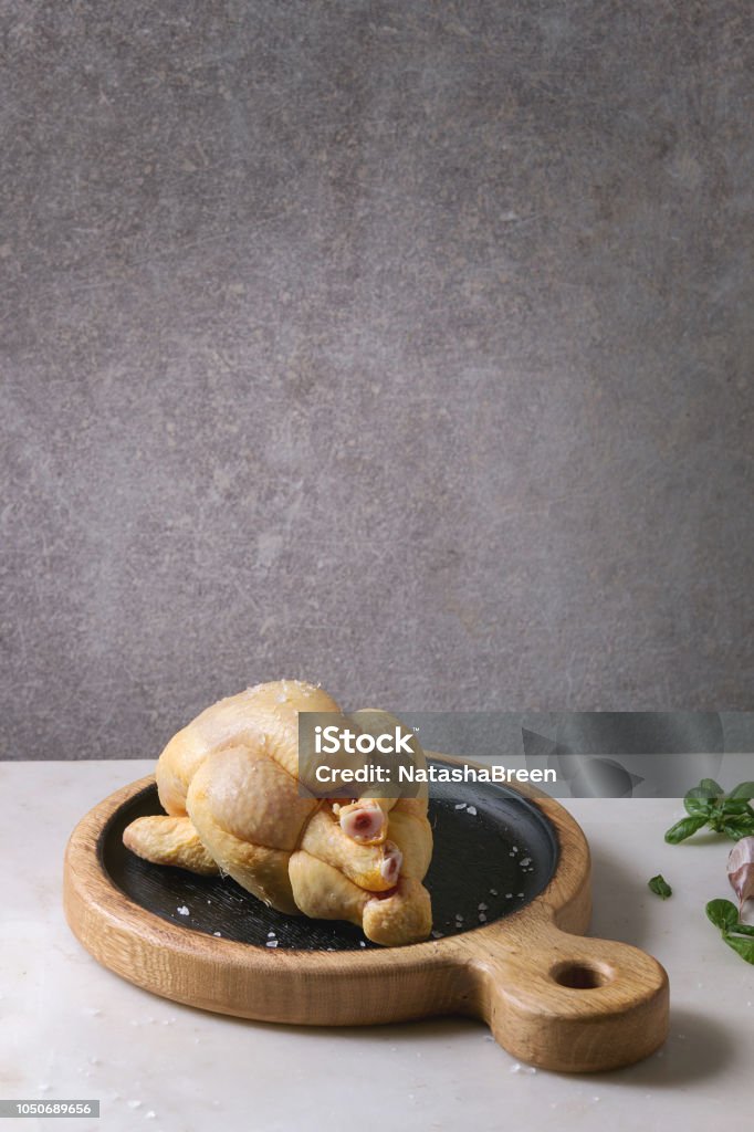 Raw uncooked chicken Raw organic uncooked whole yellow corn mini chicken on wooden cutting board with salt, herbs and garlic on white marble table with grey wall at background. Ready to cook. Animal Body Part Stock Photo