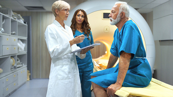 Closeup of a senior female doctor and technician preparing a senior male patient for an MRI scan of a torso. The doctor is explaining the procedure to the patient and that there's nothing to be afraid of.