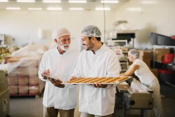 Picture of two employees in sterile clothes in food factory smiling and talking. Younger man is holding tray full of fresh cookies while the older is holding tablet and checking production line.