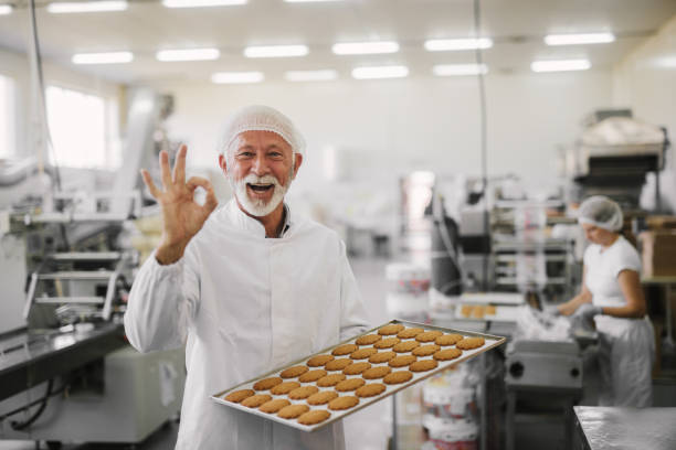 Good quality.Picture of mature cheerful male employee in sterile clothes standing in bright food factory . Holding tray full with fresh cookies and gesturing with his hand that quality is good. Good quality.Picture of mature cheerful male employee in sterile clothes standing in bright food factory . Holding tray full with fresh cookies and gesturing with his hand that quality is good. elder plant stock pictures, royalty-free photos & images