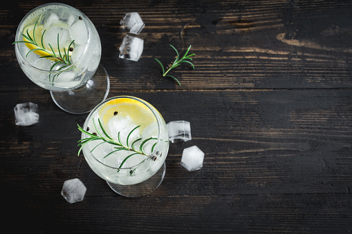 Alcohol drink (gin tonic cocktail) with lemon, rosemary and ice on rustic black wooden table, copy space, top view. Iced cocktail drink with lemon and herbs.