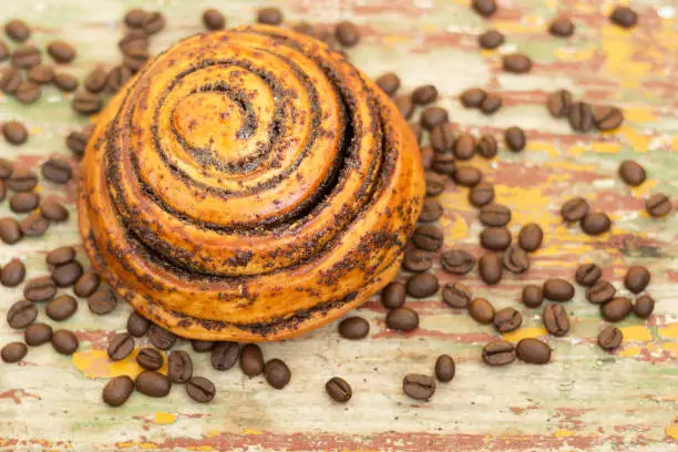 Fresh bun with poppy and cinnamon served to a cup of coffee on a wooden background. Closeup. Aromatic coffee beans. Top view.