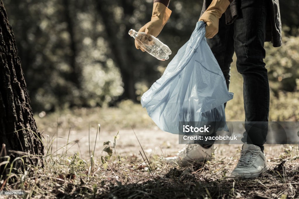 Young pupil wearing jeans and sneakers gathering empty bottles in the forest Gathering bottles. Young pupil wearing dark jeans and white sneakers gathering empty bottles in the forest Garbage Stock Photo