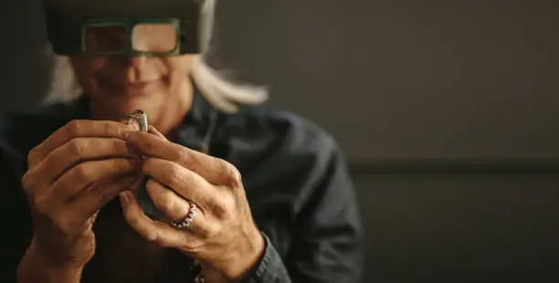 Female goldsmith inspecting jewelry using magnifying glass in workshop.  Woman jeweler inspecting diamond ring with magnifying glass.