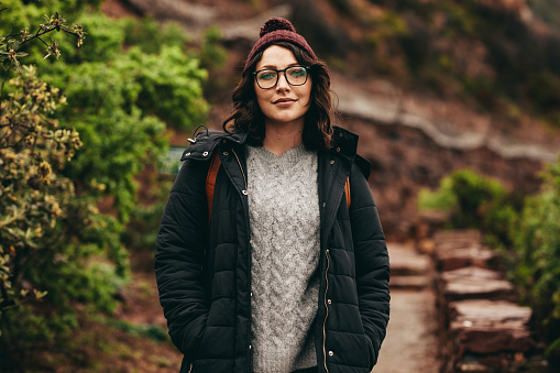 Portrait of beautiful young woman in warm clothes standing on the path at the mountain. Female tourist looking at camera standing on steps on the hill.