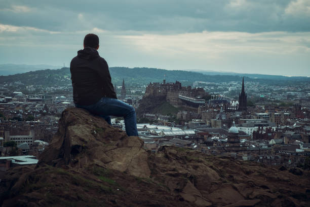 Top view of town Edinburgh. In the foreground, a man is sitting with his back looking at the city, Edinburgh, Scotland, United Kingdom
