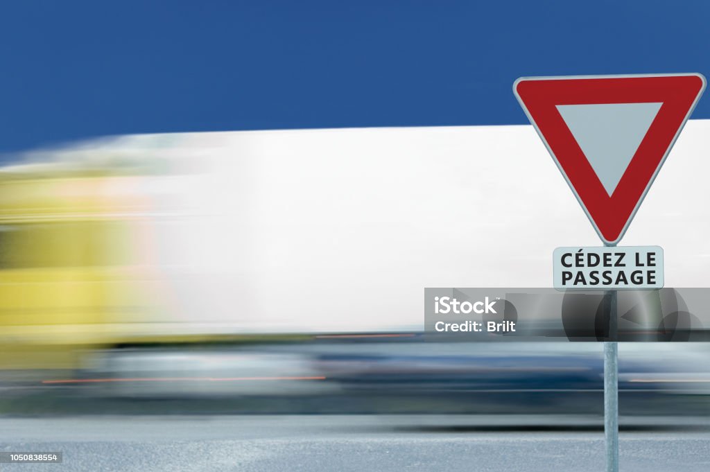 Give way yield french cédez le passage road sign, motion blurred truck vehicle traffic background, white signage triangle red frame regulatory warning, metallic pole post, blue summer sky, panneau signalisation cédez-le-passage, France Backgrounds Stock Photo