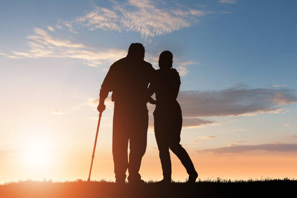 Silhouette Of Woman Assisting Her Disabled Father Silhouette Of Woman Assisting Her Disabled Father Walking In Park At Dusk custodian silhouette stock pictures, royalty-free photos & images