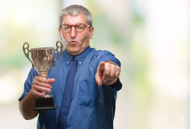 istock Handsome senior successful man holding trophy over isolated background pointing with finger to the camera and to you, hand sign, positive and confident gesture from the front 1050859002