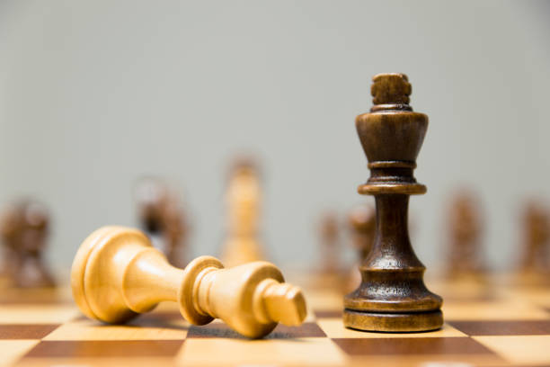 istock Two King chess pieces on a chessboard with the light king lying on its side at the foot of the victorious dark wood King 1050861652