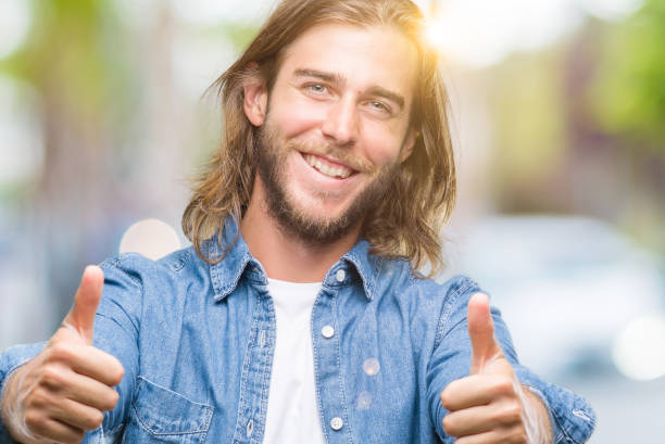 istock Young handsome man with long hair over isolated background approving doing positive gesture with hand, thumbs up smiling and happy for success. Looking at the camera, winner gesture. 1050880036