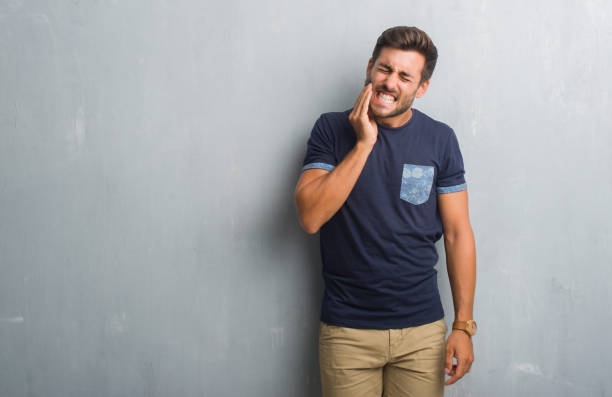 handsome young man over grey grunge wall touching mouth with hand with painful expression because of toothache or dental illness on teeth. dentist concept. - dentist pain human teeth toothache imagens e fotografias de stock