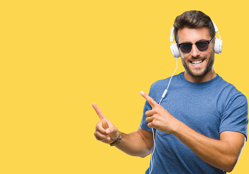 Young handsome man wearing headphones listening to music over isolated background smiling and looking at the camera pointing with two hands and fingers to the side.